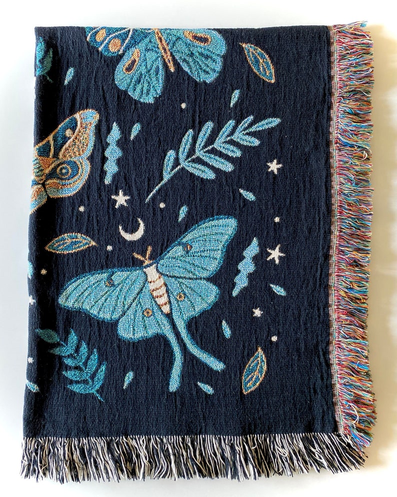 Cotton woven jacquard blanket with a fringed edge, showing the design of moths, butterflies, leaves, stars moons, and plants, folded over white background.