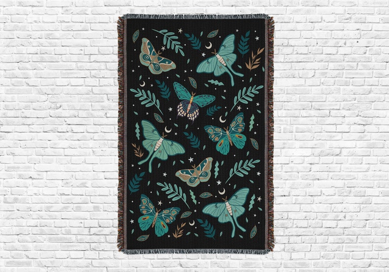 Cotton woven jacquard blanket with a fringed edge, showing the design of moths, butterflies, leaves, stars moons, and plants, full design over white brick background.