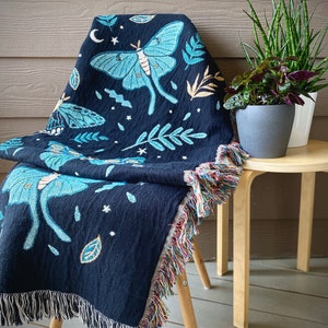 May include: A black woven throw blanket with a fringe border features a blue and green moth design with stars and leaves. The blanket is draped over a wooden chair.
