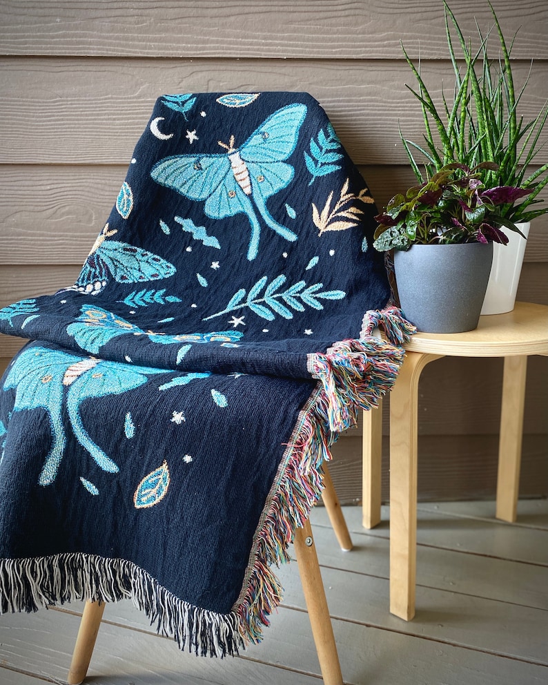 May include: A black woven throw blanket with a fringe border features a blue and green moth design with stars and leaves. The blanket is draped over a wooden chair.