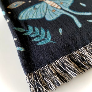 May include: Black woven throw blanket with a blue and green moth design, stars, and a crescent moon. The blanket has a fringe border.