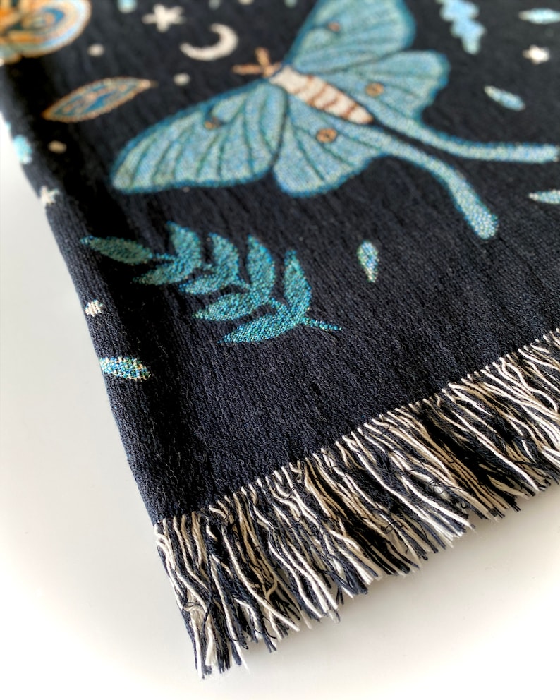 May include: Black woven throw blanket with a blue and green moth design, stars, and a crescent moon. The blanket has a fringe border.