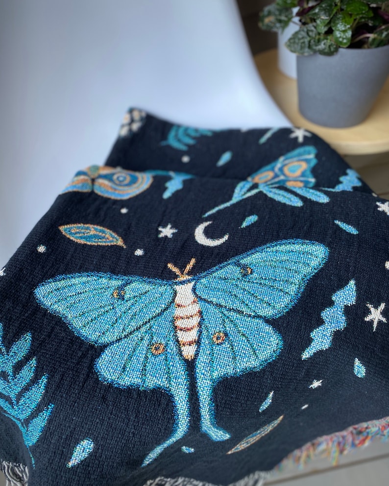 May include: A black woven tapestry throw blanket with a blue and green luna moth design. The blanket features a crescent moon, stars, and leaves.