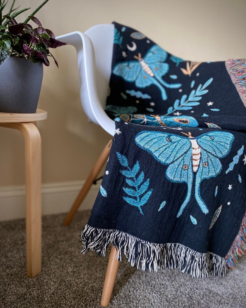 May include: A black woven throw blanket with a fringe border features a repeating pattern of blue and green moths and leaves on a dark background.
