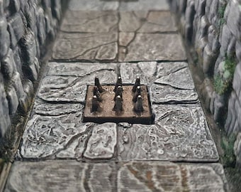 Pode incluir: Close-up de uma armadilha de masmorra em miniatura com pontas de metal que se projetam de uma base quadrada de madeira. A armadilha está colocada em um piso de pedra cinza.