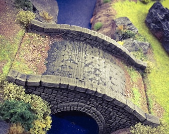 Könnte beinhalten: Eine Steinbrücke mit einem abgerundeten Bogen überspannt einen blauen Bach. Die Brücke ist aus grauem Stein und hat eine raue Textur. Der Bach ist von grünem Gras und brauner Erde umgeben.