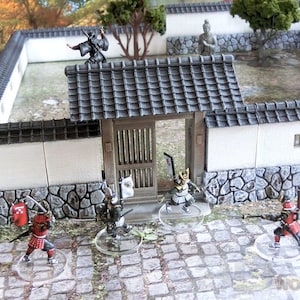 May include: A miniature Japanese garden scene with a white wall, a gate, and several figures. The figures are painted in black, red, and gold. The scene is set on a gray stone path.