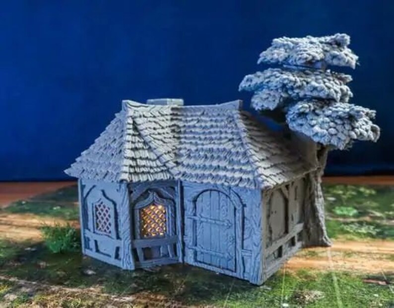 Puede incluir: Una caba&ntilde;a en miniatura gris con un techo de paja y un peque&ntilde;o &aacute;rbol que crece en un lado. La caba&ntilde;a tiene dos ventanas y una puerta. Las ventanas est&aacute;n iluminadas con un brillo c&aacute;lido.