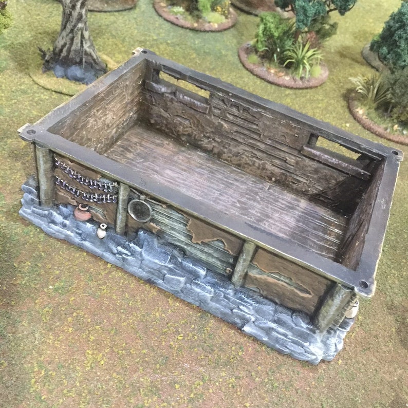 Op de afbeelding: Een houten miniatuurgebouw met een stenen fundering. Het gebouw heeft een houten dak en muren met een klein raam. De binnenkant is leeg en het gebouw is klaar voor gebruik in een tabletop-spel of diorama.