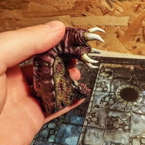 May include: A hand holds a small, painted, pink and brown monster figurine with white claws. The figurine has a textured surface and appears to be made of plastic.