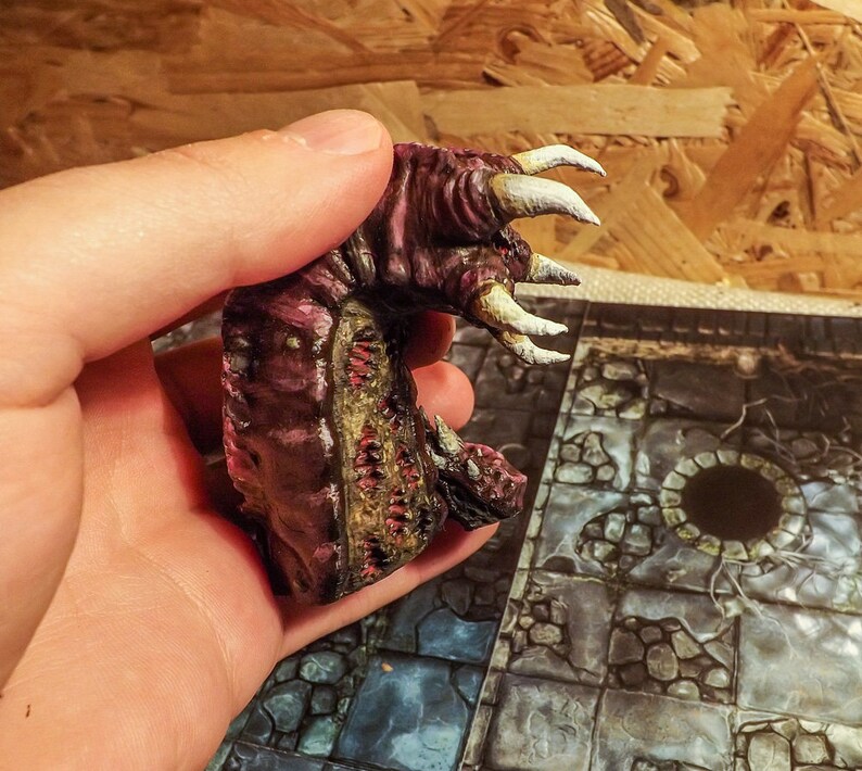 May include: A hand holds a small, painted, pink and brown monster figurine with white claws. The figurine has a textured surface and appears to be made of plastic.
