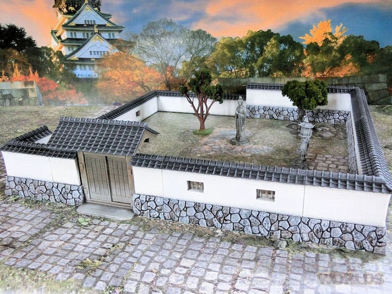 May include: A miniature white walled Japanese garden with a small gray roofed building, a stone path, and two small trees.