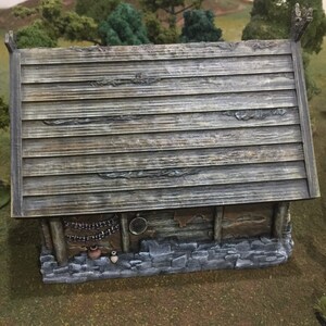 Op de afbeelding: Een klein houten huis met een verweerd grijs dak en een stenen fundering. Het huis heeft een klein raam en een deur. Er staan twee kleine potten op de grond voor het huis.