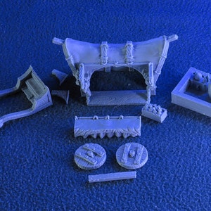 May include: A grey plastic model kit of a medieval market stall with a canvas awning, wooden frame, and various accessories. The kit includes a cart, barrels, and other items.
