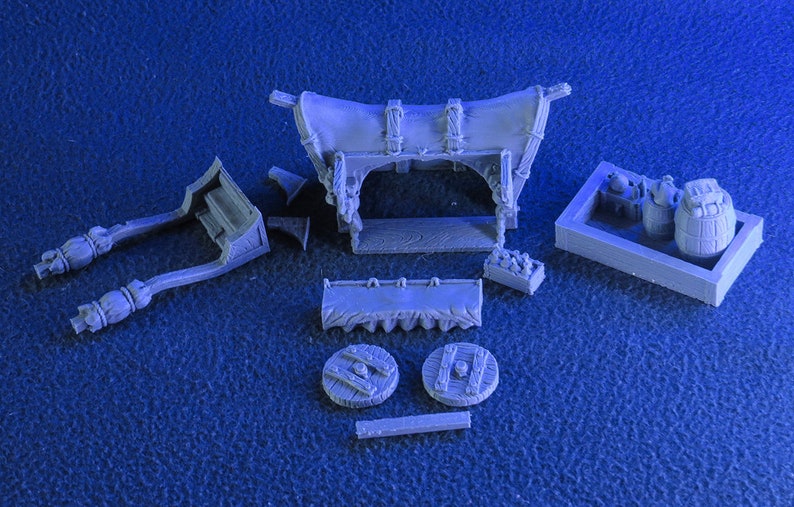 May include: A grey plastic model kit of a medieval market stall with a canvas awning, wooden frame, and various accessories. The kit includes a cart, barrels, and other items.