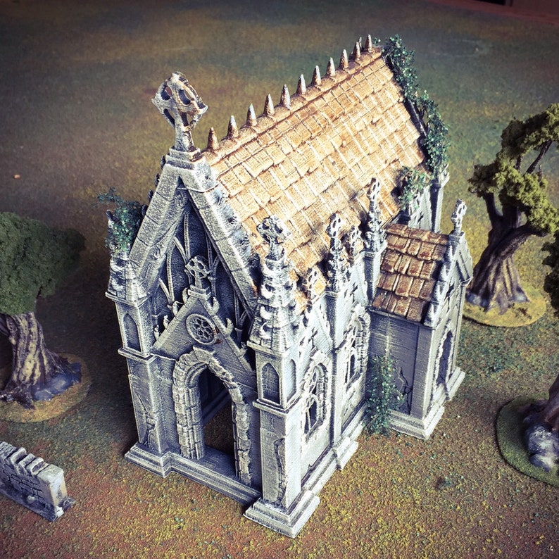 May include: A miniature stone chapel with a brown tiled roof and ivy growing on the walls. The chapel has a pointed arch doorway and several windows. The chapel is painted in a light gray color.