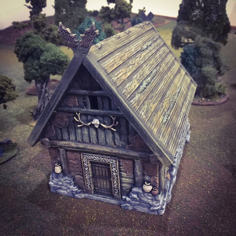 Op de afbeelding: Een klein houten huis met een zadeldak en een stenen fundering. Het huis heeft een deur en twee ramen, en er zijn twee geweistangen boven de deur gemonteerd. Het dak is bedekt met houten shingles en heeft een decoratieve houten nok.