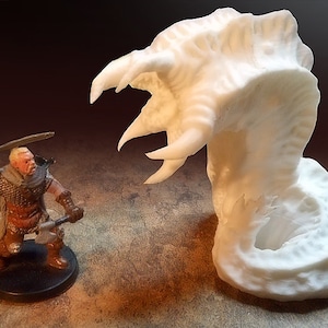 May include: A white 3D printed miniature of a monster with a large, open mouth and sharp teeth. The monster is facing a miniature of a warrior holding a sword and an axe.