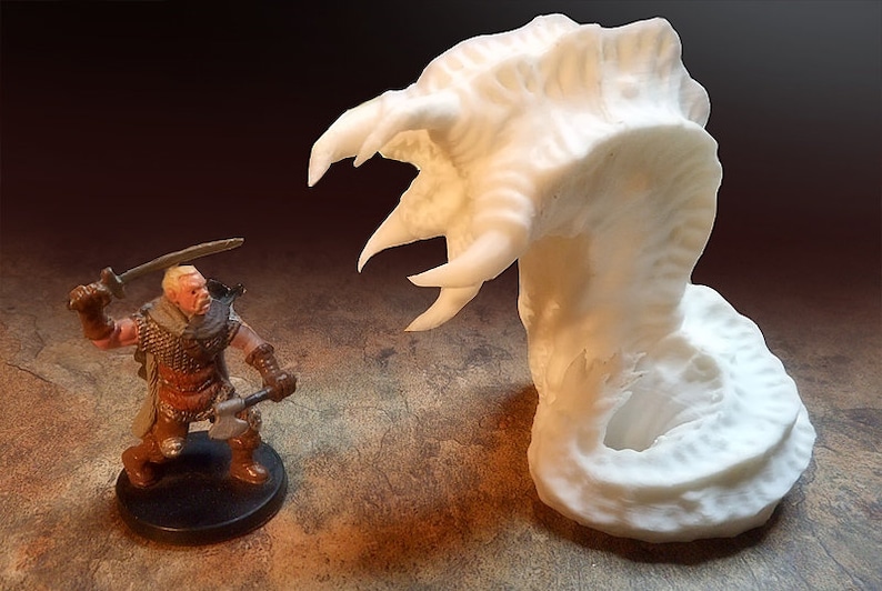 May include: A white 3D printed miniature of a monster with a large, open mouth and sharp teeth. The monster is facing a miniature of a warrior holding a sword and an axe.