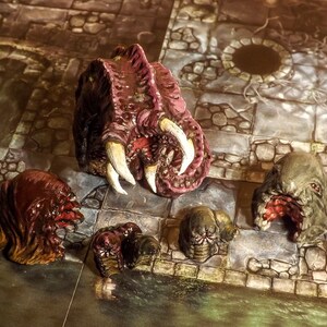 May include: Five miniature painted figures of grotesque creatures with open mouths and sharp teeth. The figures are made of a dark brown, red, and green material and are set on a stone-like surface.