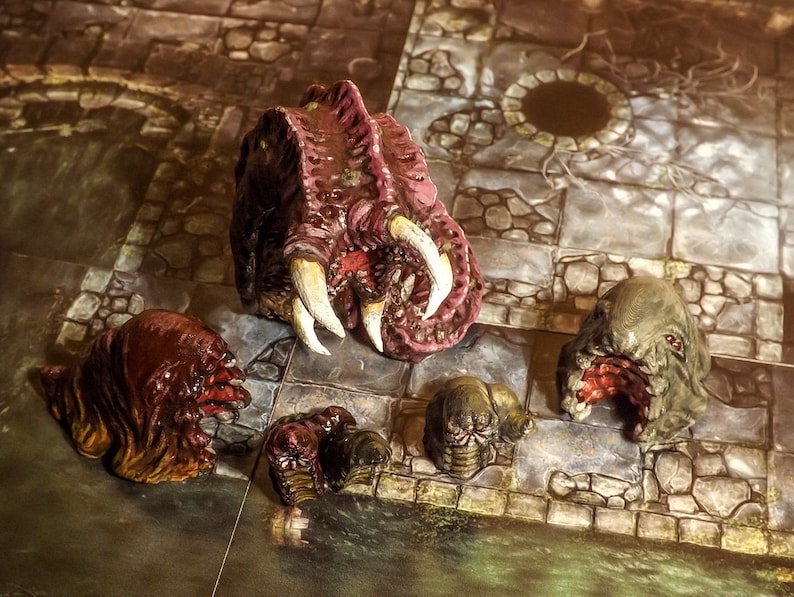 May include: Five miniature painted figures of grotesque creatures with open mouths and sharp teeth. The figures are made of a dark brown, red, and green material and are set on a stone-like surface.