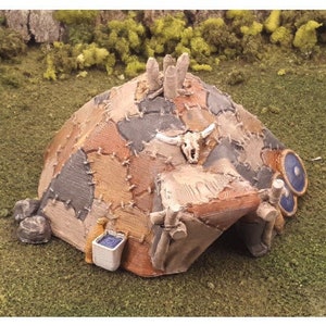 Puede incluir: Un modelo en miniatura de una caba&ntilde;a r&uacute;stica remendada con una puerta de madera y una decoraci&oacute;n de calavera. La caba&ntilde;a est&aacute; hecha de materiales marrones, grises y blancos y tiene un barril azul y blanco en el lateral.