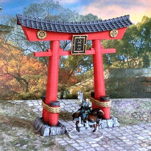 May include: A red and black miniature torii gate with a gold trim and a sign with Japanese characters. The gate is set on a stone base with rope accents. A miniature samurai figure on horseback is in front of the gate.