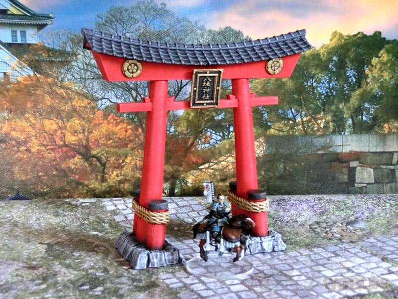 May include: A red and black miniature torii gate with a gold trim and a sign with Japanese characters. The gate is set on a stone base with rope accents. A miniature samurai figure on horseback is in front of the gate.