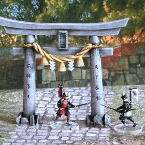 May include: Two miniature samurai figures stand facing each other in front of a traditional Japanese torii gate. The gate is made of gray stone and has a gold rope and bells hanging from it. The figures are painted in black and red armor and hold swords.