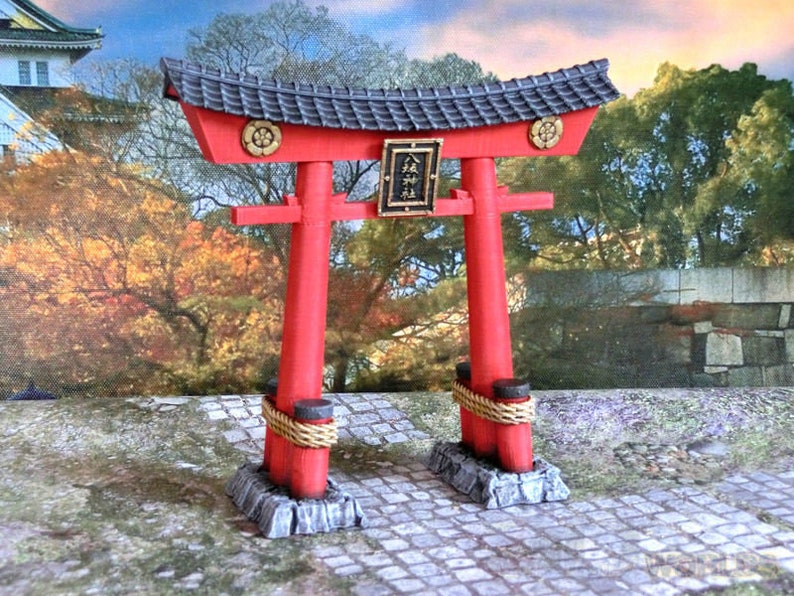 May include: A red and black miniature torii gate with a gold plaque that reads "松" in Japanese. The gate is set on a stone base and surrounded by a cobblestone path.