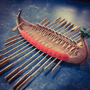 May include: A wooden model of a Viking longship with red shields along the sides and wooden oars. The ship has a dragon head at the bow.