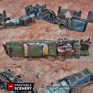 May include: A green and yellow rusted car wreck with a pink interior is partially visible behind a fence made of wood and metal. The car is surrounded by scrap metal and other debris. The scene is a model for a tabletop game or diorama.