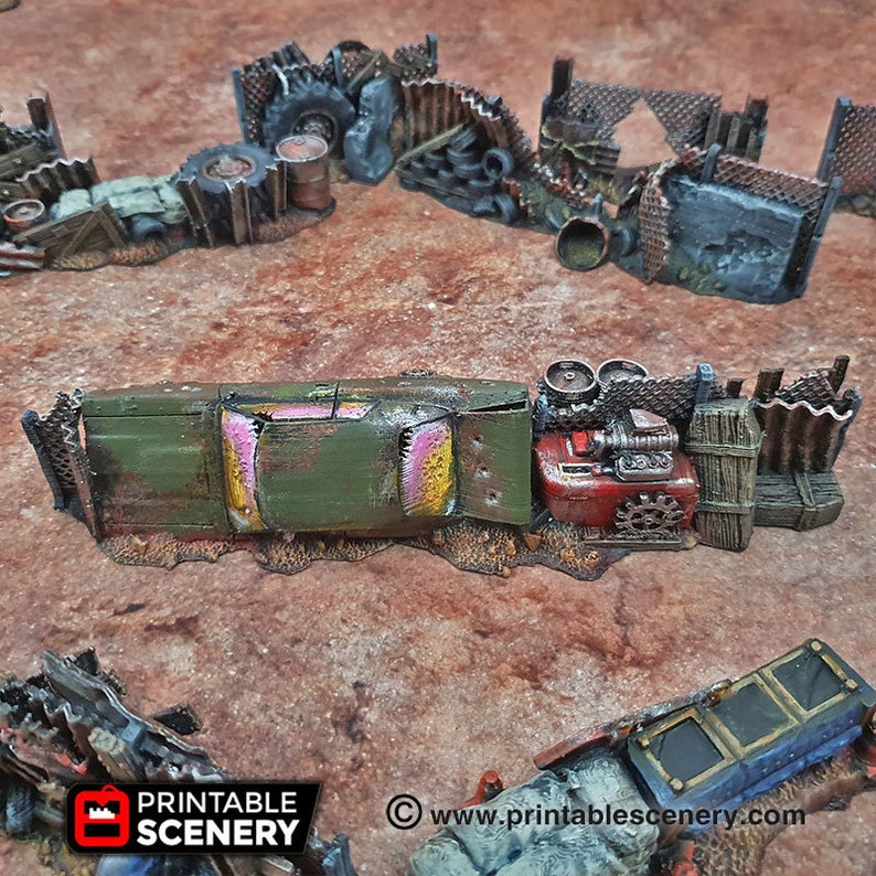 May include: A green and yellow rusted car wreck with a pink interior is partially visible behind a fence made of wood and metal. The car is surrounded by scrap metal and other debris. The scene is a model for a tabletop game or diorama.
