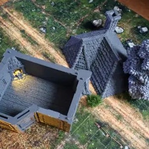 Puede incluir: Dos edificios en miniatura pintados de gris, uno con techo y otro con interior abierto, sobre una superficie texturizada verde y marr&oacute;n.