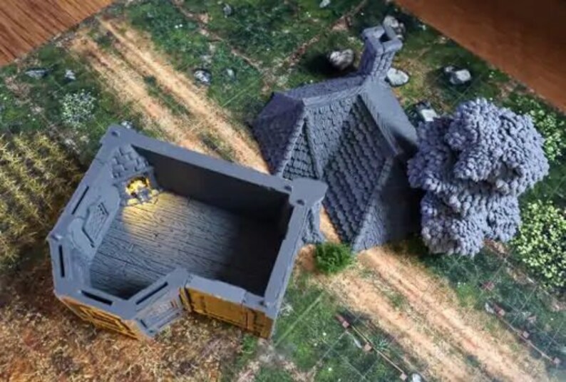 Puede incluir: Dos edificios en miniatura pintados de gris, uno con techo y otro con interior abierto, sobre una superficie texturizada verde y marr&oacute;n.