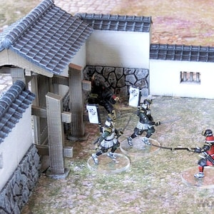 May include: A miniature scene depicting a battle between samurai warriors. The scene features a Japanese-style gate, a wall, and several figures in traditional samurai armor. The figures are holding swords and are engaged in combat. The scene is painted in a realistic style and is suitable for use in tabletop wargaming.
