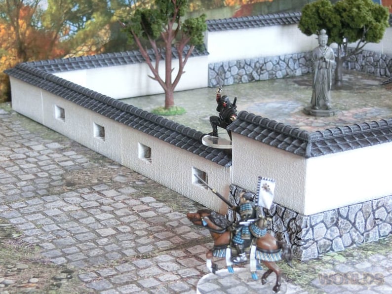 May include: A miniature scene depicting a Japanese garden with a white wall, a stone path, and a small tree. Two figures, one in black and one in armor, are positioned near the wall. The scene is designed for tabletop gaming.