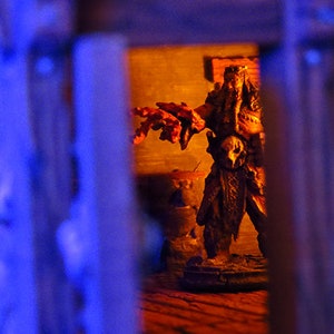 Puede incluir: Primer plano de una figura en miniatura de color marr&oacute;n oscuro y detallada de una criatura con una m&aacute;scara de calavera, de pie en una habitaci&oacute;n tenue con paredes de color naranja. La figura sostiene un arma en su mano derecha y tiene una larga barba fluida.