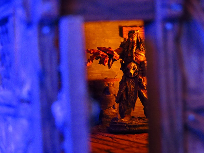 Puede incluir: Primer plano de una figura en miniatura de color marr&oacute;n oscuro y detallada de una criatura con una m&aacute;scara de calavera, de pie en una habitaci&oacute;n tenue con paredes de color naranja. La figura sostiene un arma en su mano derecha y tiene una larga barba fluida.