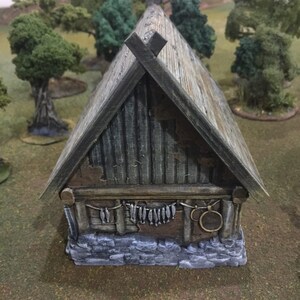 Op de afbeelding: Een klein, verweerd, grijs en bruin, miniatuurhuisje van hout met een zadeldak en een stenen fundering. Het huisje heeft een houten deur en ramen, en een rij vissen die aan de voorkant hangen.