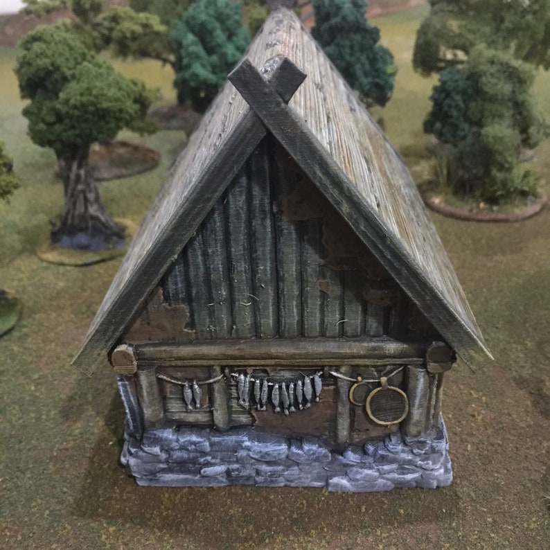 Op de afbeelding: Een klein, verweerd, grijs en bruin, miniatuurhuisje van hout met een zadeldak en een stenen fundering. Het huisje heeft een houten deur en ramen, en een rij vissen die aan de voorkant hangen.