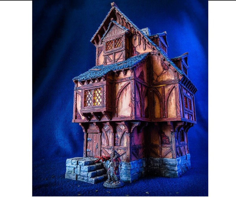 Puede incluir: Un modelo en miniatura detallado de un edificio de tres pisos con estructura de madera y cimientos de piedra. El edificio tiene un techo de inclinaci&oacute;n pronunciada, varias ventanas y una peque&ntilde;a puerta. El modelo est&aacute; pintado en tonos de marr&oacute;n, gris y blanco, y se coloca sobre un fondo azul.