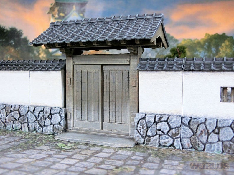 May include: A miniature Japanese-style gate with a gray tiled roof and wooden doors. The gate is set into a white wall with a stone base.