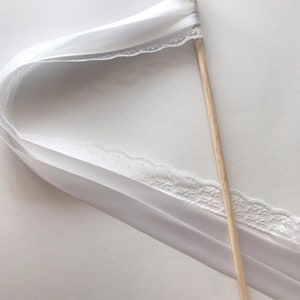 May include: White satin ribbon wand with a gold bell at the top. The ribbon has a lace trim and is attached to a wooden stick.