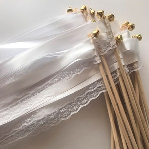 May include: White satin and lace ribbon wands with gold bells attached to wooden sticks. The wands are tied together in a bundle.