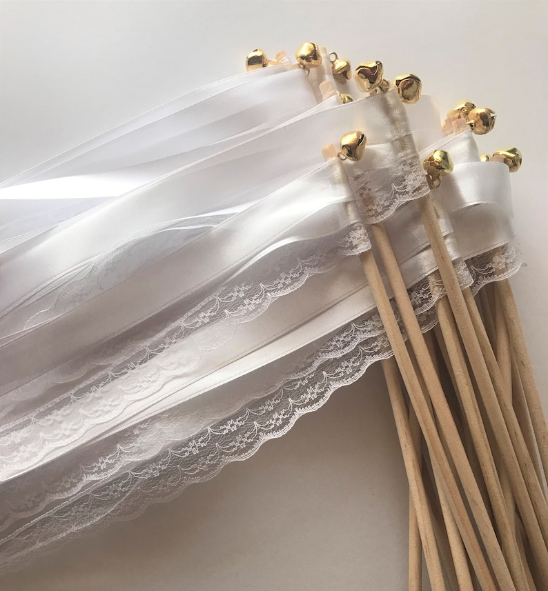 May include: White satin and lace ribbon wands with gold bells attached to wooden sticks. The wands are tied together in a bundle.