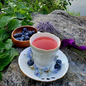 May include: A white teacup with a pink beverage sits on a white saucer with blue flowers and blueberries. A wooden bowl filled with blueberries sits next to the teacup. A bouquet of lavender with a purple ribbon is in the background.
