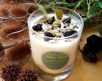 May include: A white candle in a glass jar with a green label that reads "Garden of Moonligh Dark Moon Cleansing & Banishing All Natural Gemstone & Herbs Hand Poured With Love & A Dash of Magic". The candle is surrounded by dried plants and black stones.