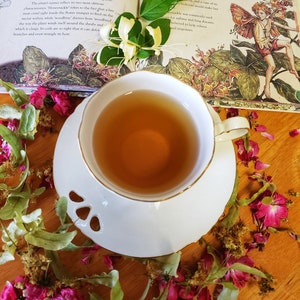 May include: A white teacup with a saucer filled with tea sits on a wooden surface surrounded by pink and green flower petals. The teacup has a gold rim. The background is a book with a floral illustration and text about honeysuckle.