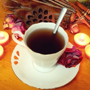 May include: A white teacup and saucer with a gold rim, filled with dark tea and a silver spoon. The teacup is on a wooden table with a dried flower, candles, and other decorative items.